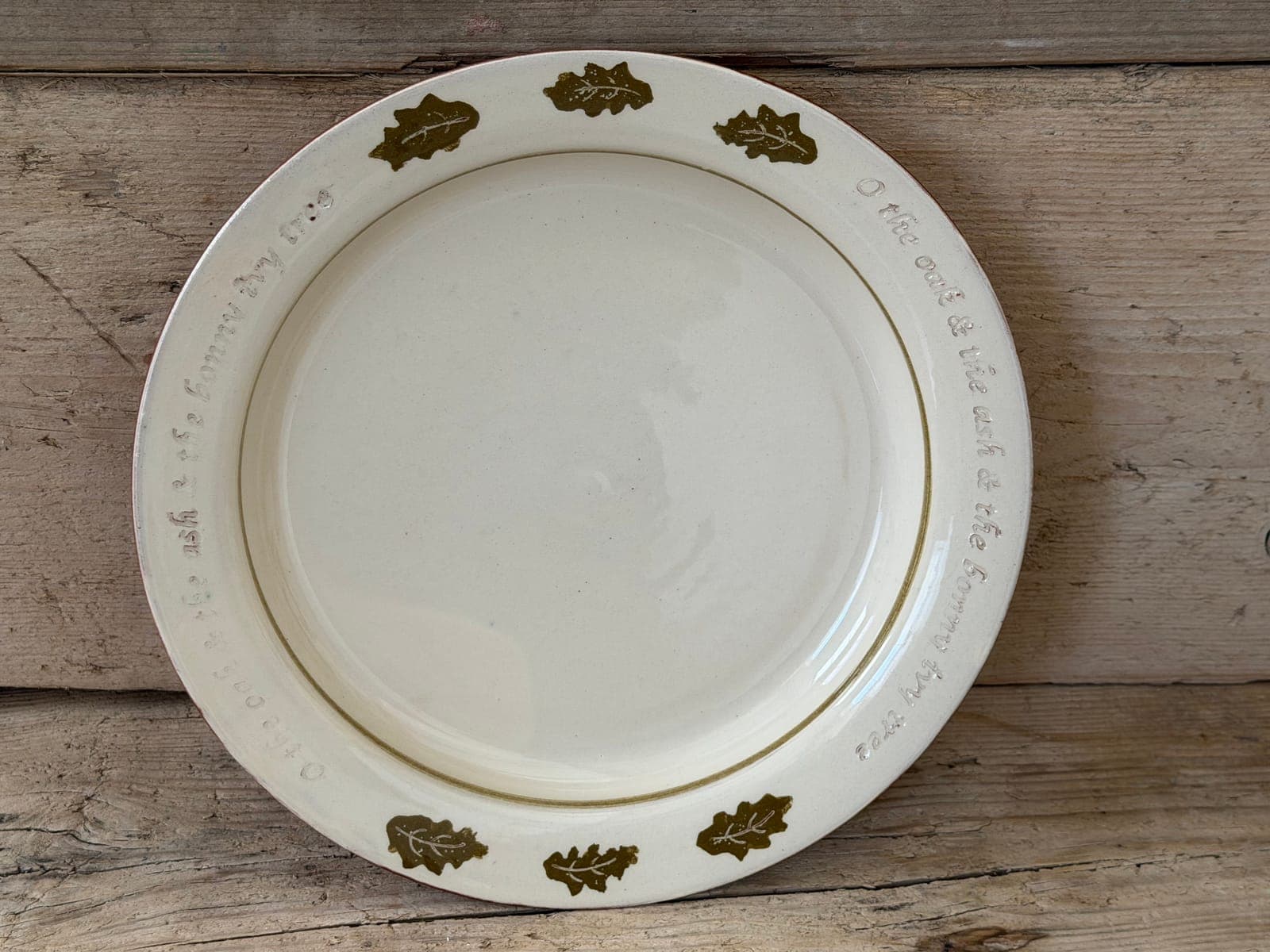 Oak Leaf slipware plate in cream slip from above, rim decorated with olive oak leaves and an inscribed folk-song verse, on rustic wood