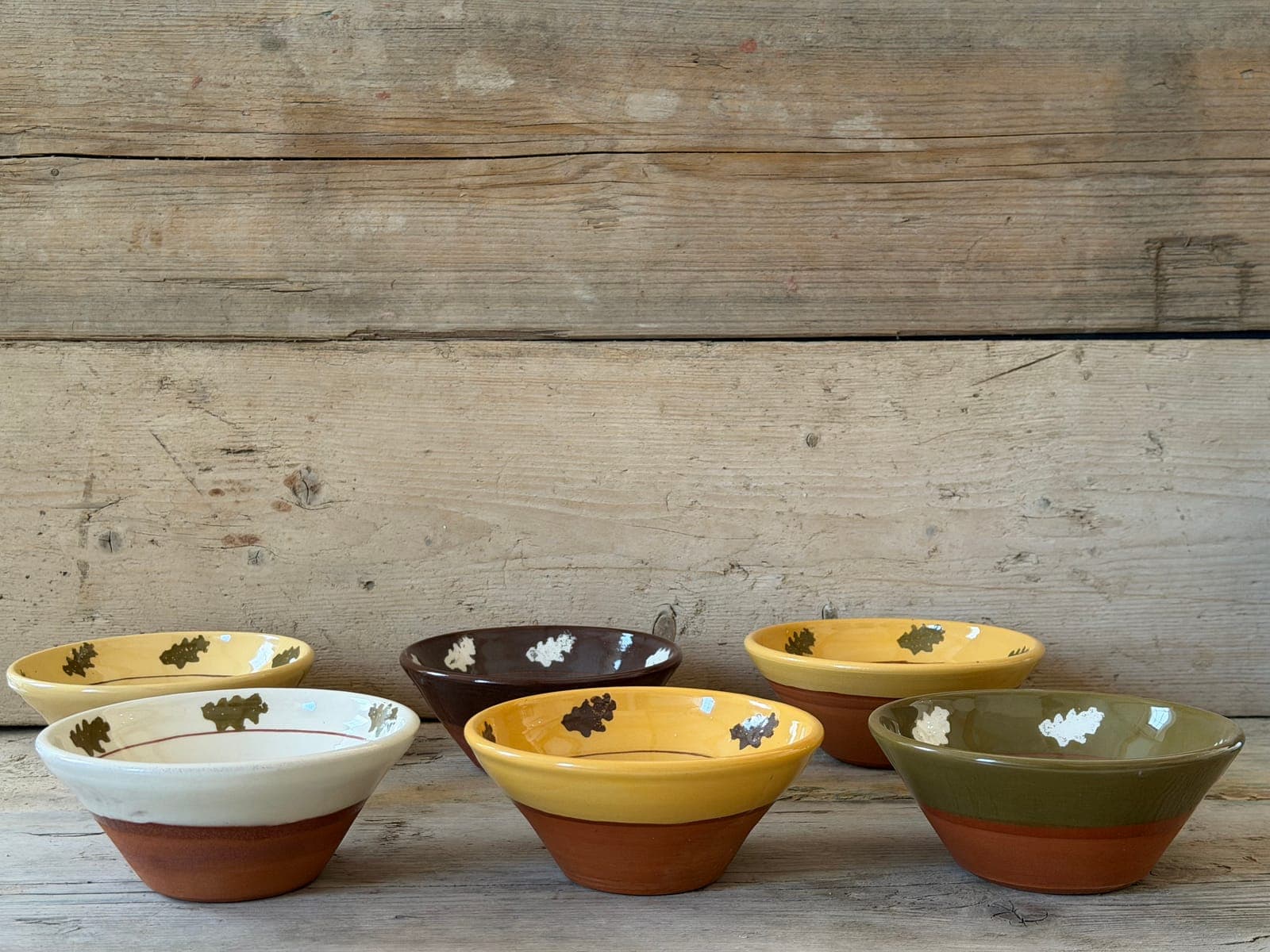 Group of six Oak Leaf slipware small bowls in cream, mustard, olive and chestnut slips with oak-leaf motifs, arranged on rustic wooden boards