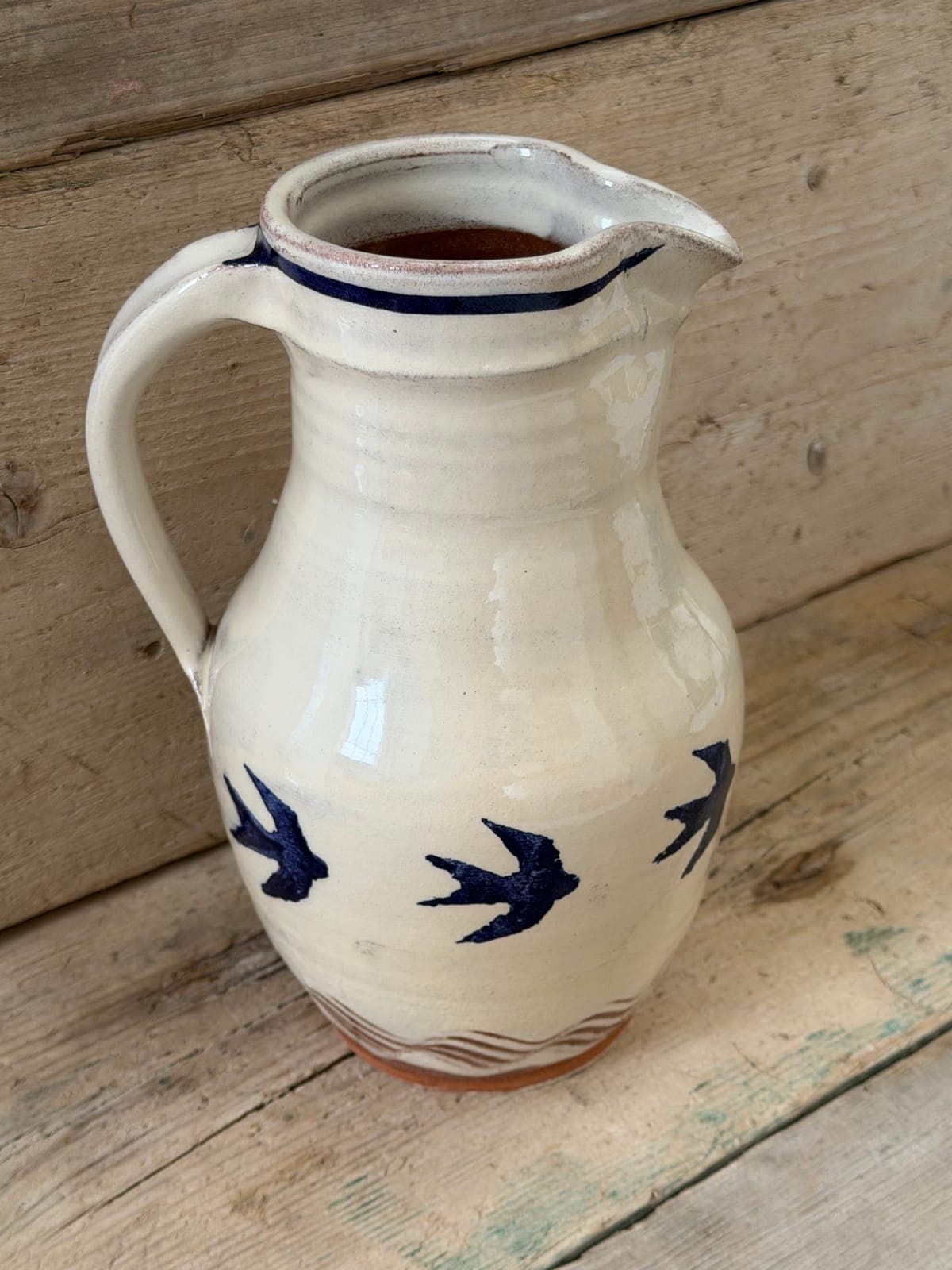 Swallows slipware jug standing upright with the whole handle in frame, cream glaze with dark blue swallows and terracotta wave combing at the base